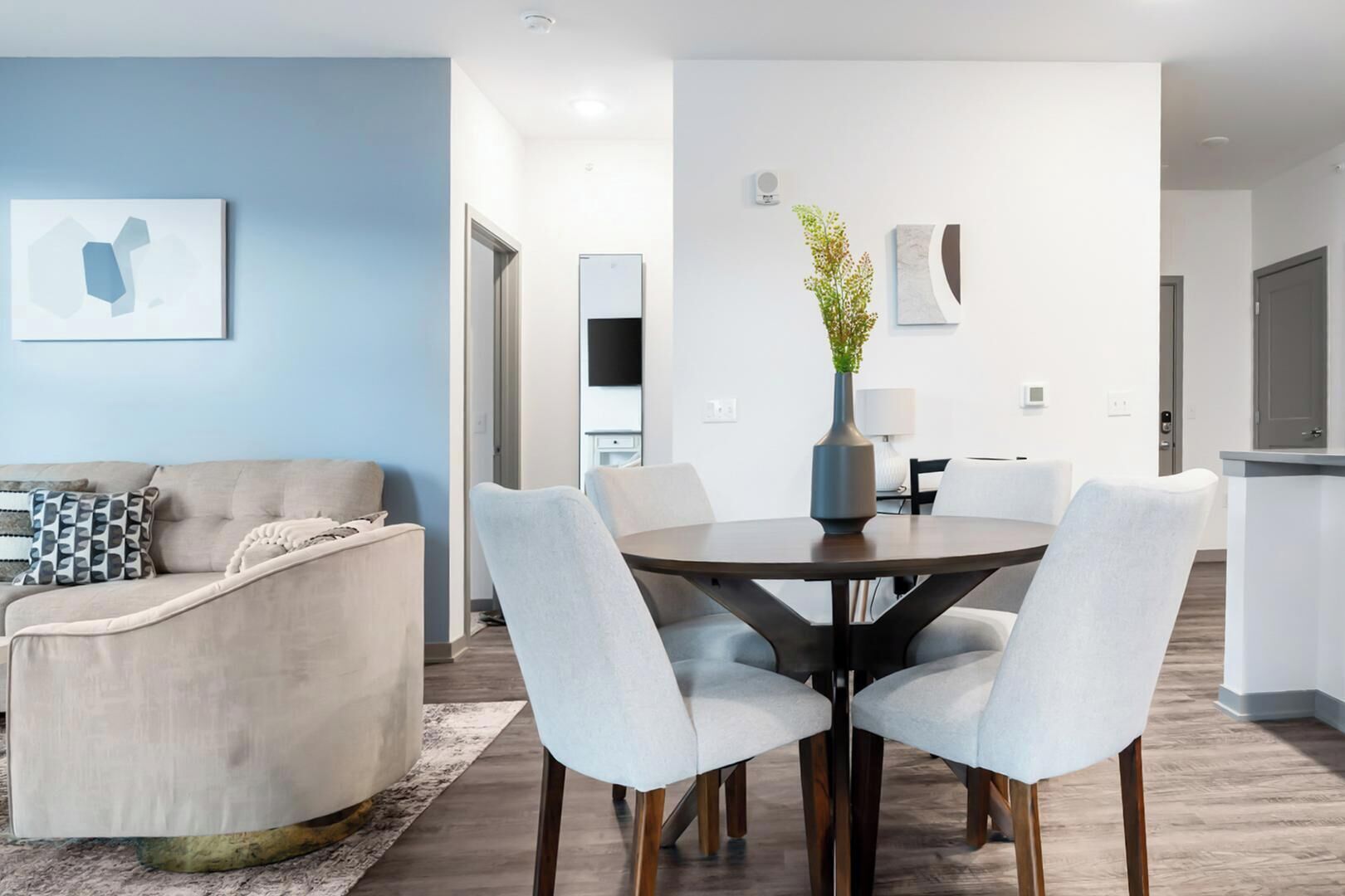 Un salon moderne avec un canapé, une table à manger et des chaises, un vase avec une plante, et un coin cuisine à l’arrière-plan.
