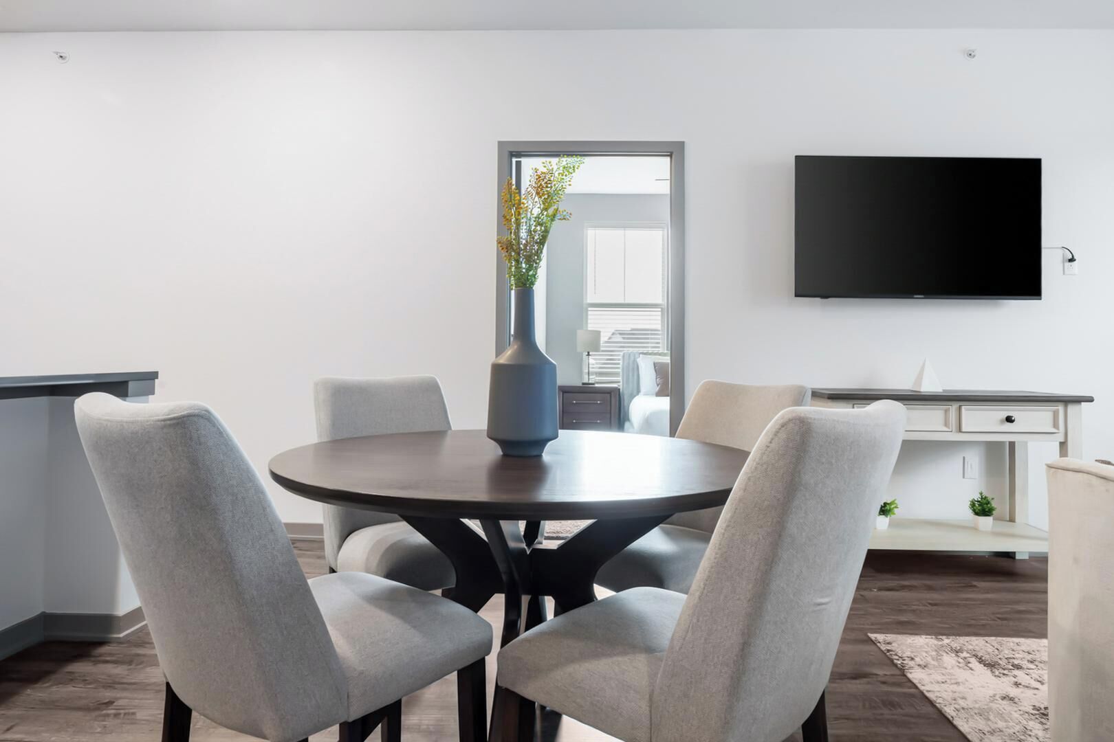 A modern dining area with a round table, four grey chairs, a vase with flowers, a flat-screen TV on the wall, and a white cabinet.