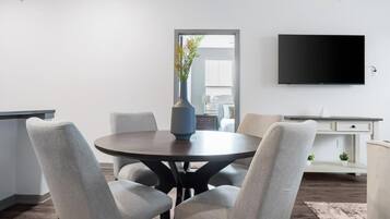 A modern dining area with a round table, four grey chairs, a vase with flowers, a flat-screen TV on the wall, and a white cabinet.