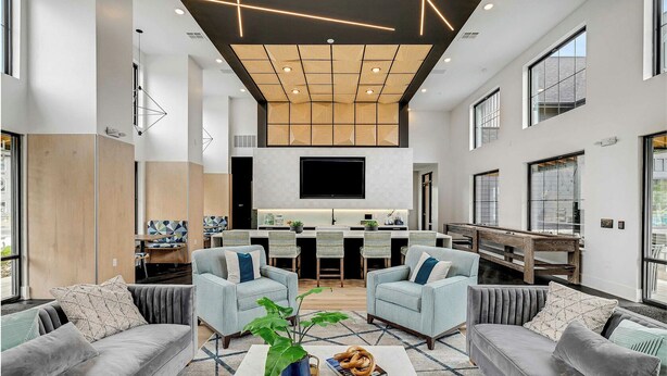 Ein modernes Wohnzimmer mit grauen Sofas, einem Couchtisch mit Pflanze und einer Schüssel Brezeln sowie eine Küchenzeile mit Fernseher über der Arbeitsplatte.