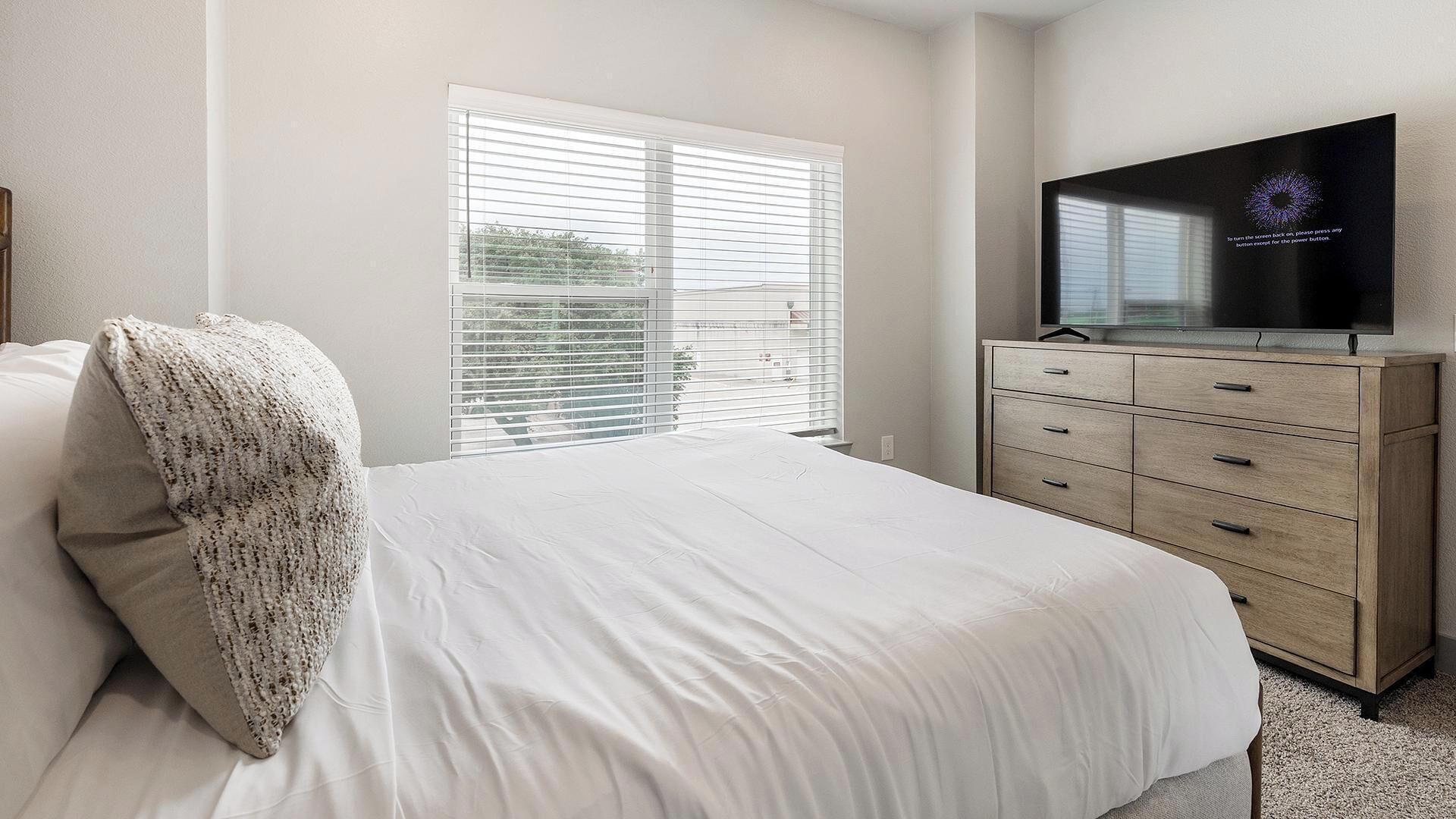 Een slaapkamer met een bed, een televisie op een houten dressoir en een raam met jaloezieën.