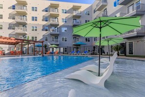 Ein Schwimmbecken mit Liegestühlen und Sonnenschirmen, umgeben von einem mehrstöckigen Gebäude mit Balkonen.