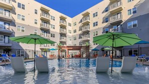 A pool area with lounge chairs and umbrellas, surrounded by a multi-story building with balconies. - Landing | Chic 2BD in Grand Prairie (Grand Prairie)