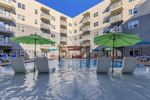 A pool area with lounge chairs and umbrellas, surrounded by a multi-story building with balconies. - Landing | Chic 2BD in Grand Prairie (Grand Prairie)