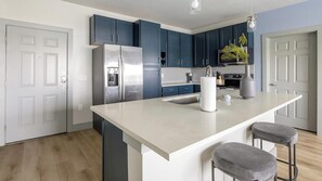 A modern kitchen with dark blue cabinets, a stainless steel refrigerator, a white countertop, and two bar stools. - Landing | Modern 2BD in Grand Prairie (Grand Prairie)