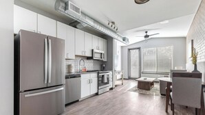 A modern kitchen with stainless steel appliances, a dining area with a table and chairs, and a large window with blinds. - Landing | Stylish 1BD, Pool, Rooftop Lounge (Bellview)