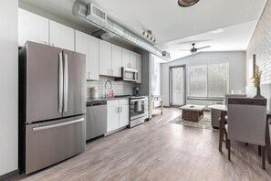 A modern kitchen with stainless steel appliances, a dining area with a table and chairs, and a large window with blinds. - Landing | Modern 1BD, Pool, Gym (Bellview)