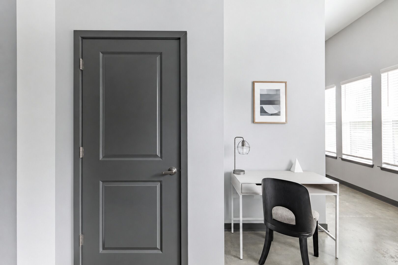 A modern office space with a closed door, a desk with a chair, a framed picture on the wall, and large windows.