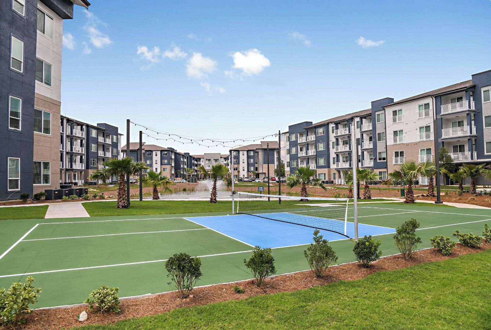 Una cancha deportiva con una red, rodeada de un área ajardinada con árboles pequeños y una fuente de agua, con edificios residenciales de varios pisos al fondo.