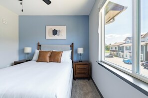 Ein Schlafzimmer mit einem Bett, zwei Nachttischen, einem Fenster mit Blick auf Gebäude und einem an der Wand hängenden abstrakten Kunstwerk.