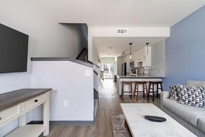A modern kitchen with a central island, a dining area with bar stools, a sofa with patterned cushions, and a flat-screen TV mounted on the wall. - Landing | Beautiful 2BD, Yoga Studio, Gym (Bellview)