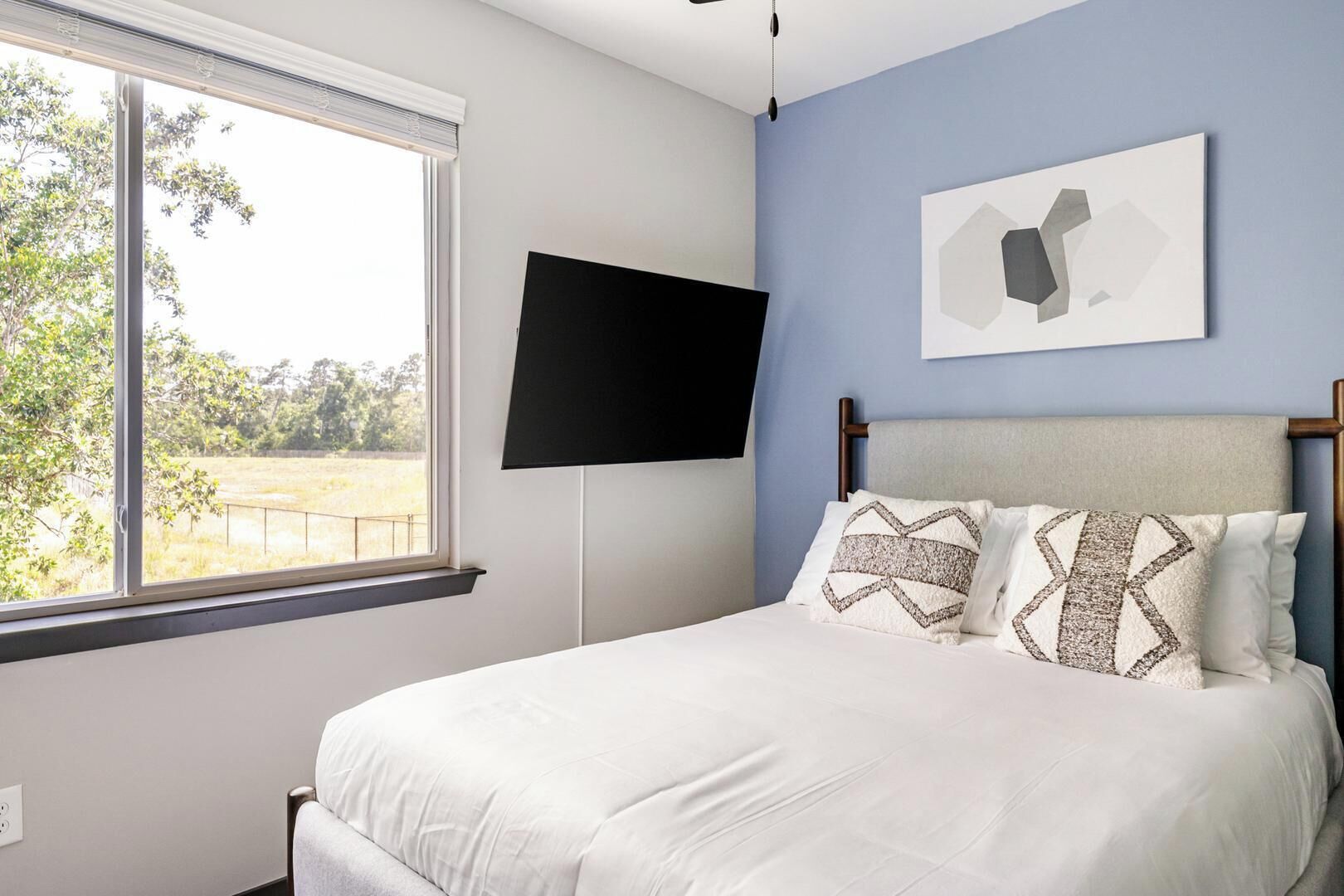 A bedroom with a bed, a TV mounted on the wall, and a window with a view of trees and grass.