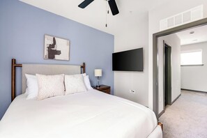 A bedroom with a bed, a nightstand, a lamp, a painting, a TV, and a door leading to another room.