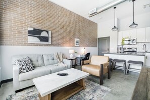 Een moderne woonkamer met een bank, salontafel en een stoel. Er is een keuken met een fornuis en kasten.