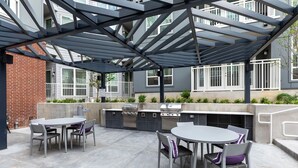 Een terras buiten met een barbecue en zithoek, omgeven door een pergola van metaal.
