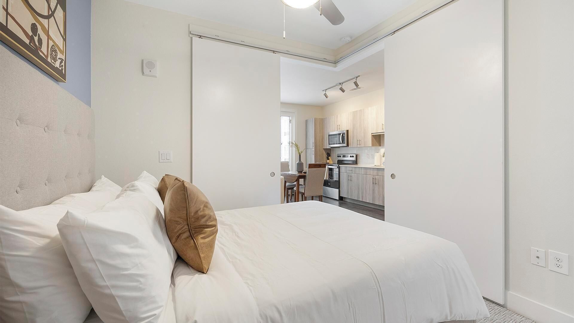Een slaapkamer met een bed, kussens, een hoofdbord en een deur die leidt naar een keuken.