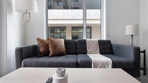 A modern living room with a grey sofa, a white coffee table, and a view of a building through the window. - Landing | Modern 1BD, Gym (Austin)