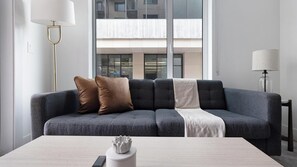 A modern living room with a grey sofa, a white coffee table, and a view of a building through the window. - Landing | Modern 1BD, Gym (Austin)