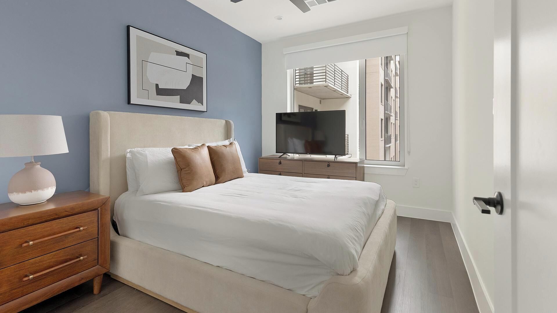 A bedroom with a bed, a bedside table, a lamp, a TV, and a window with a view of buildings.