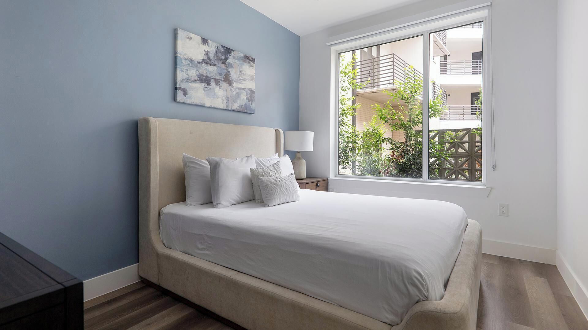 A bedroom with a bed, a nightstand, a lamp, a window with a view of buildings and greenery, and a painting on the wall.