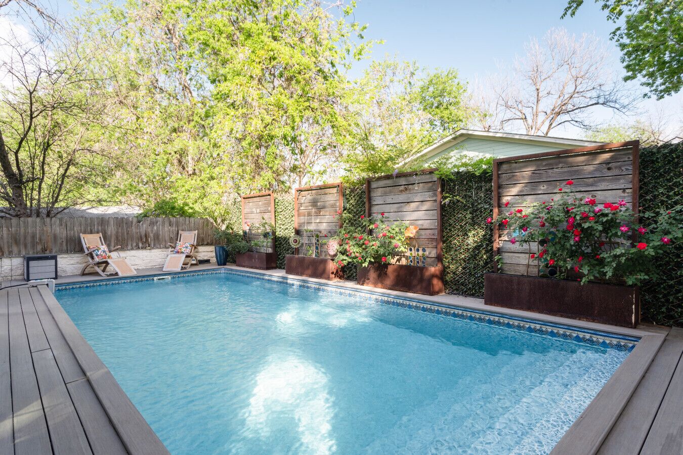 A swimming pool with wooden deck and lounge chairs, surrounded by a wooden fence and flowering plants.