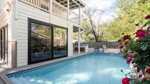 Une piscine avec une terrasse en bois, un patio couvert et un bâtiment blanc doté de grandes fenêtres.