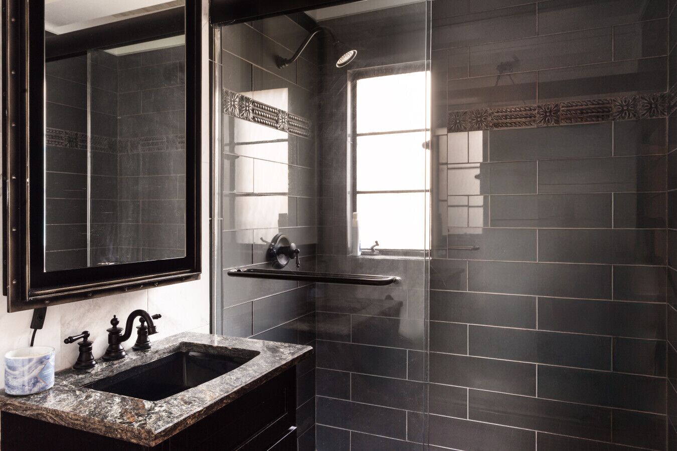 A bathroom with a dark tiled wall, a large mirror, a sink with two faucets, and a window.