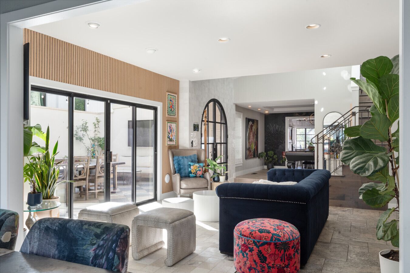 Un vaste espace de vie avec une porte-fenêtre coulissante donnant sur une terrasse extérieure, un canapé bleu foncé, un fauteuil beige et un pouf rouge.
