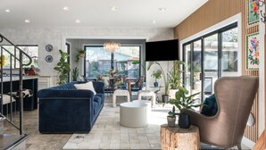 Un salon spacieux avec un canapé bleu, une table basse ronde et un fauteuil marron. Un escalier mène à un autre niveau, et il y a un coin repas avec une table et des chaises.