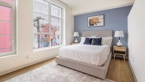 Een slaapkamer met een bed, twee nachtkastjes met lampen, een raam met zonwering en een muur met een ingelijst kunstwerk.