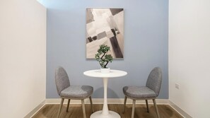 Ein Zimmer mit einem runden weißen Tisch, zwei grau gepolsterten Stühlen, einem kleinen Topfpflanzen und einem großen abstrakten Gemälde an der Wand.