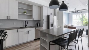 Une cuisine moderne avec des meubles en bois clair, un réfrigérateur en acier inoxydable, un îlot central intégré avec des tabourets, et des luminaires suspendus.