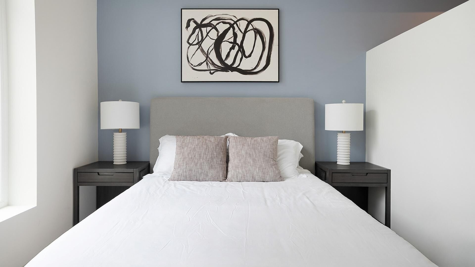 Une chambre moderne avec un grand lit, deux tables de chevet avec des lampes, une tête de lit grise et une œuvre d’art abstraite accrochée au mur.