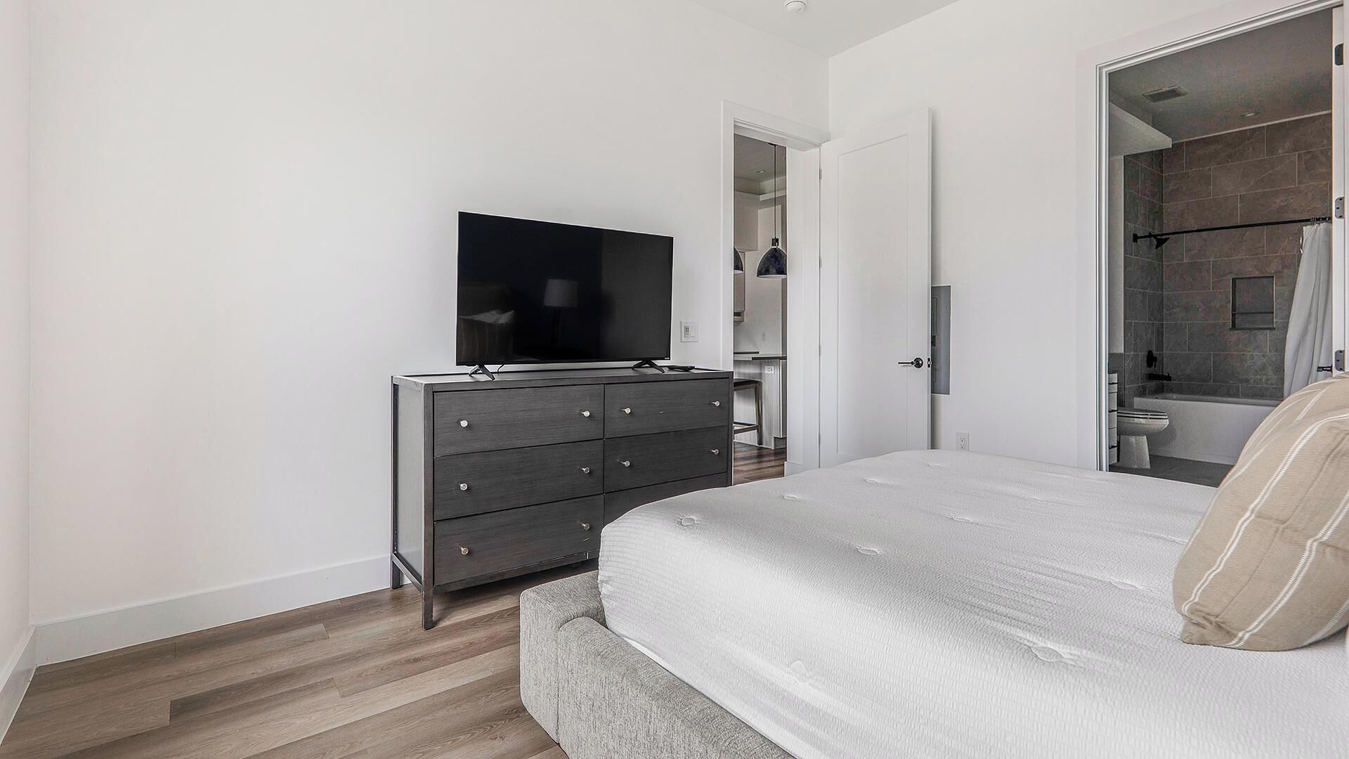 A bedroom with a bed, a dresser, a television, and a bathroom visible through an open door.
