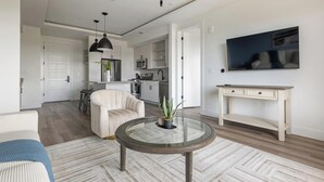 Ein modernes Wohnzimmer mit einem Glastisch, einem beigen Sessel, einem Essbereich mit Barhocker und einer Küche mit weißen Schränken.