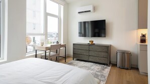 Una habitación de hotel moderna con cama, escritorio con silla, televisor montado en la pared, un mueble pequeño y un cubo de basura.