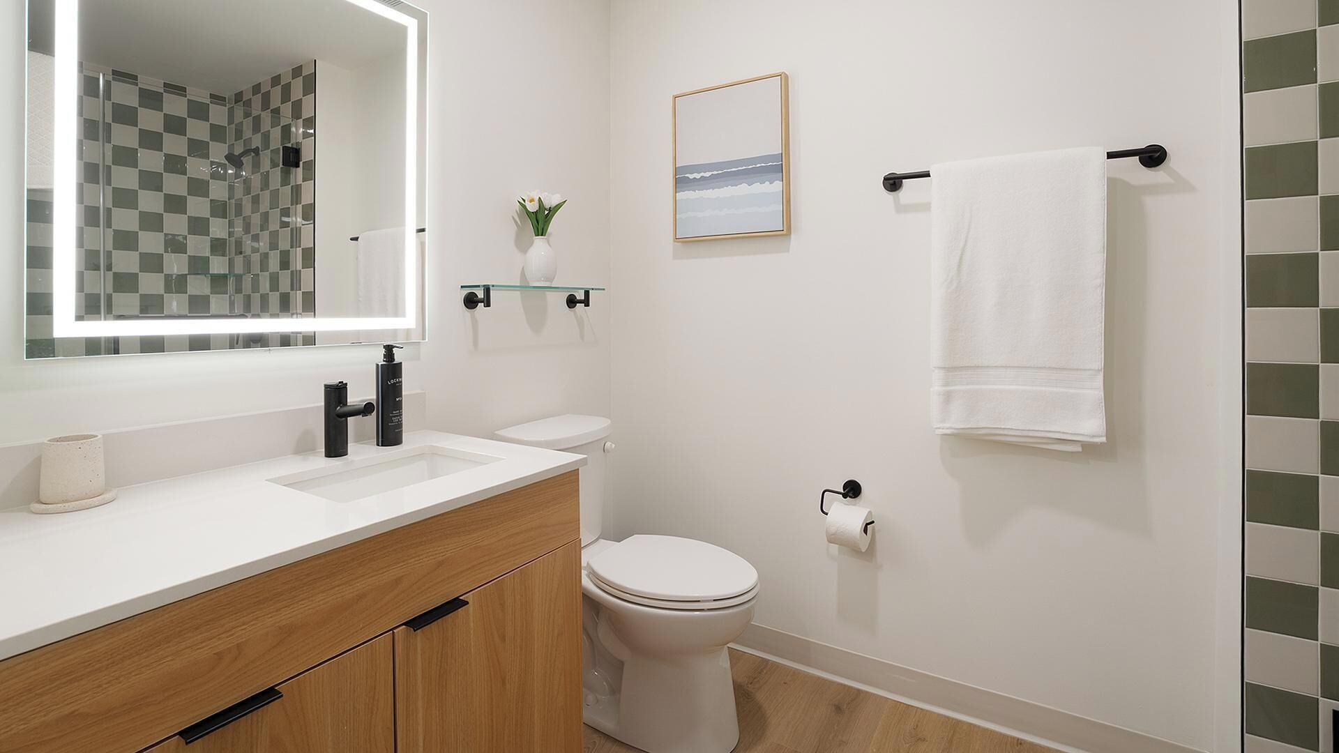 Une salle de bain moderne avec un mur de douche à carreaux, des toilettes blanches, un lavabo avec un miroir et un tableau encadré accroché au mur.