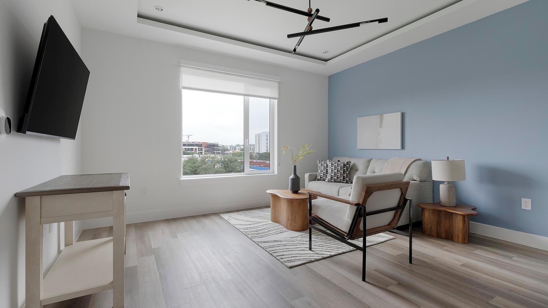 Un salon moderne avec un canapé, une table basse, un téléviseur fixé au mur et une grande fenêtre donnant sur des immeubles.
