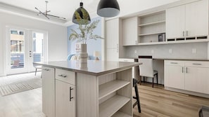 Eine moderne Küche mit hellen Holzschränken, einer zentralen Kochinsel mit einer Vase, Pendelleuchten und Blick ins Freie.