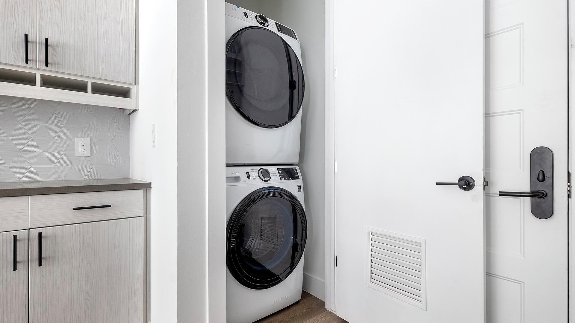 A laundry room with two stacked front-loading washing machines, a wooden cabinet with drawers, and a door with a handle.