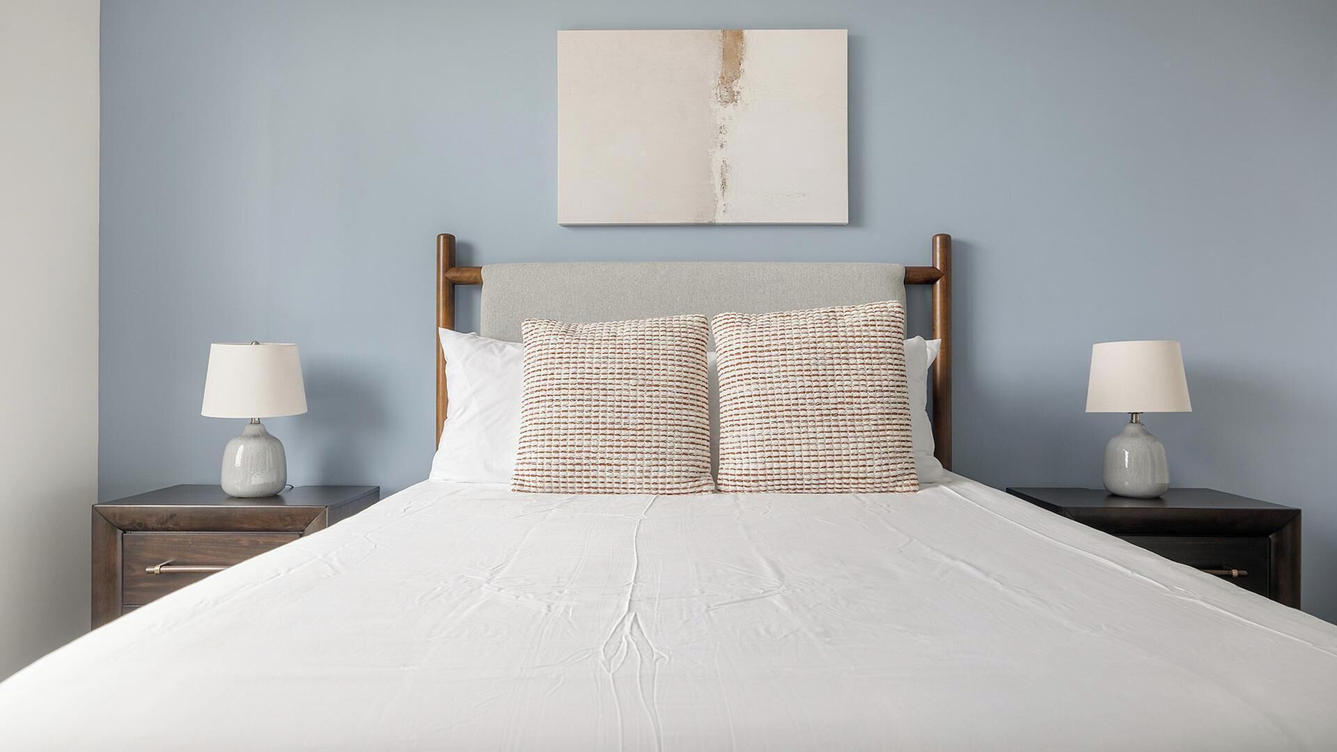 A bed with a wooden headboard, two bedside tables with lamps, and a plain wall with a single piece of artwork above the bed.