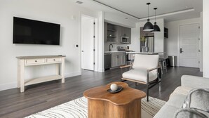 Uma sala de estar moderna com uma TV de tela plana, uma mesa de centro de madeira, uma poltrona branca e uma cozinha ao fundo.