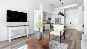 Een moderne woonkamer met een flatscreen televisie, een houten salontafel, een vaas met bloemen, een witte fauteuil en een keuken op de achtergrond.