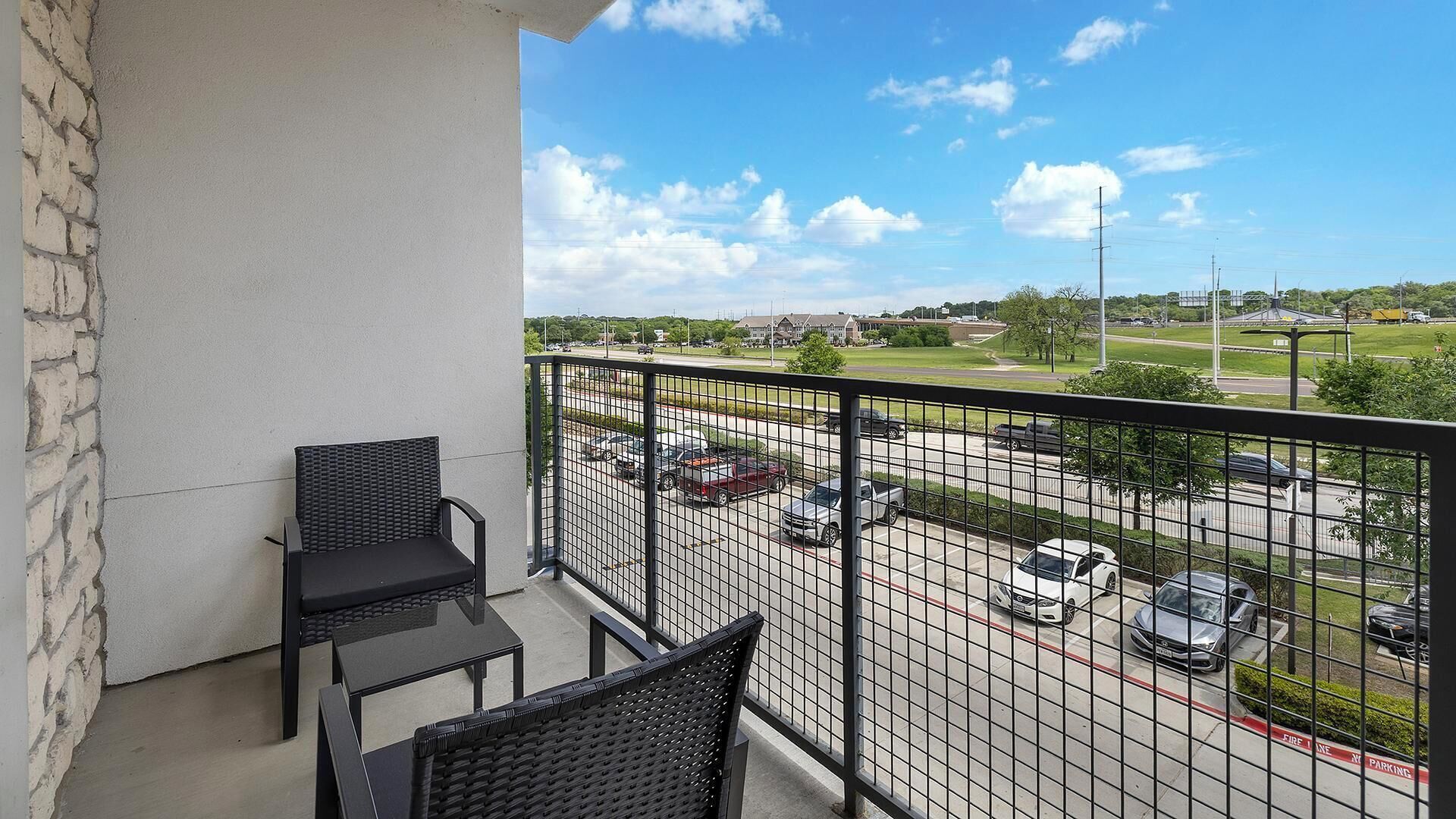 A balcony with two chairs and a table, overlooking a parking lot with several cars and a building in the background.