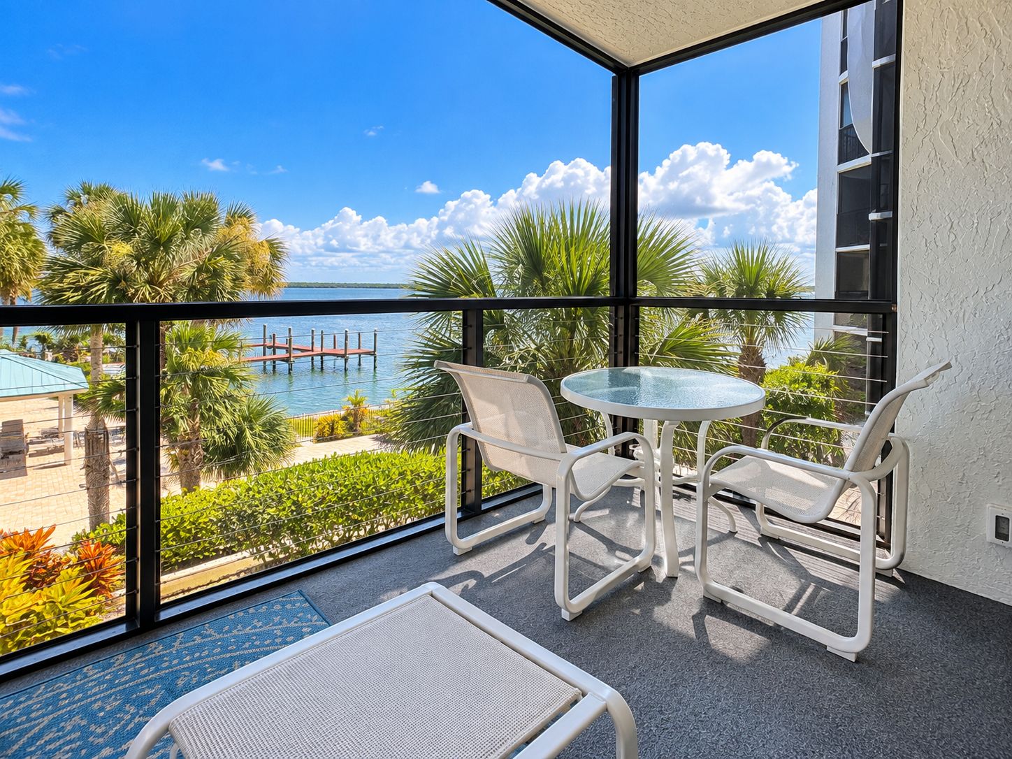 A balcony with a glass table and two chairs, overlooking a marina with boats and palm trees.
