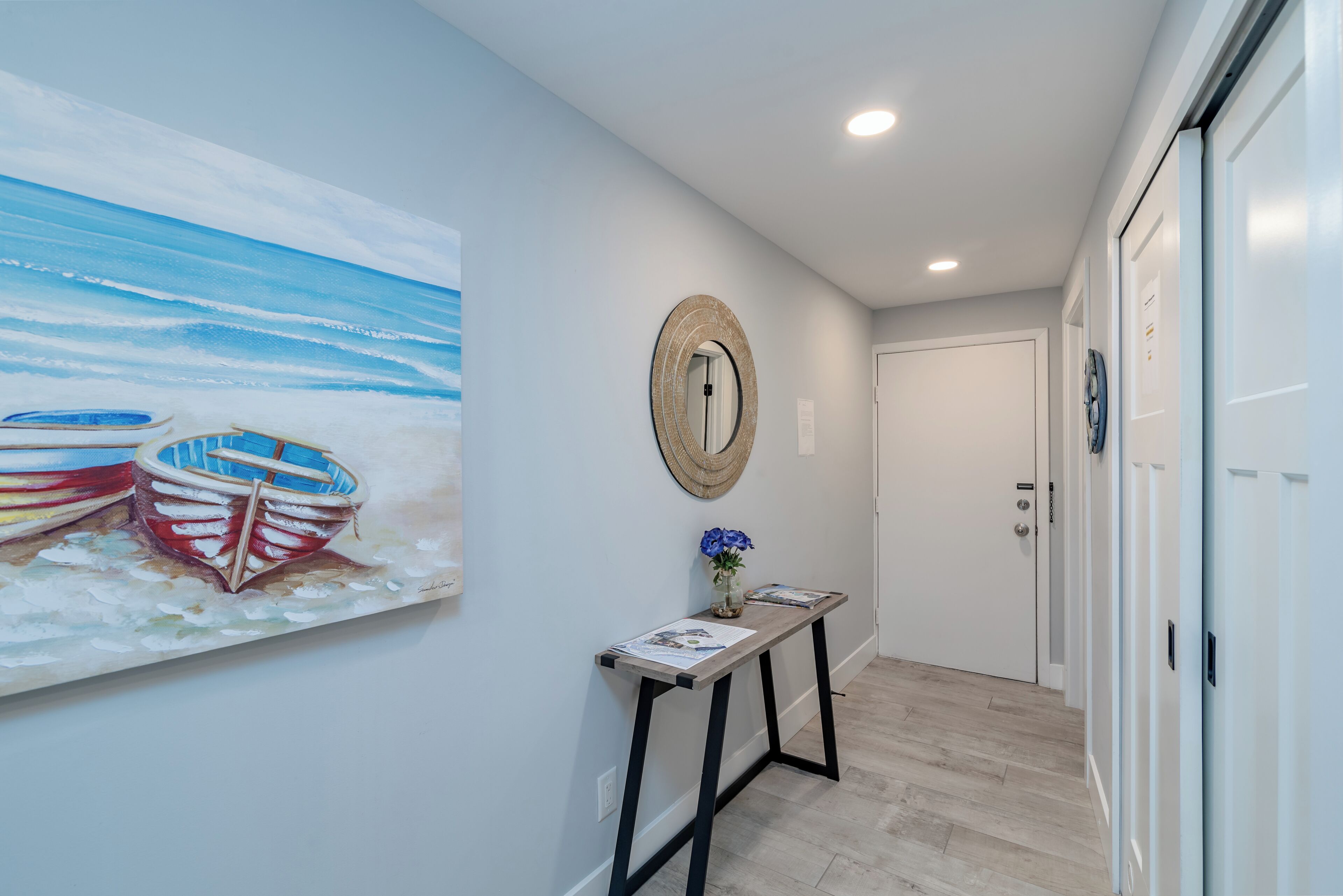 A hallway with a painting of boats, a round mirror, and a small table with a vase of flowers.