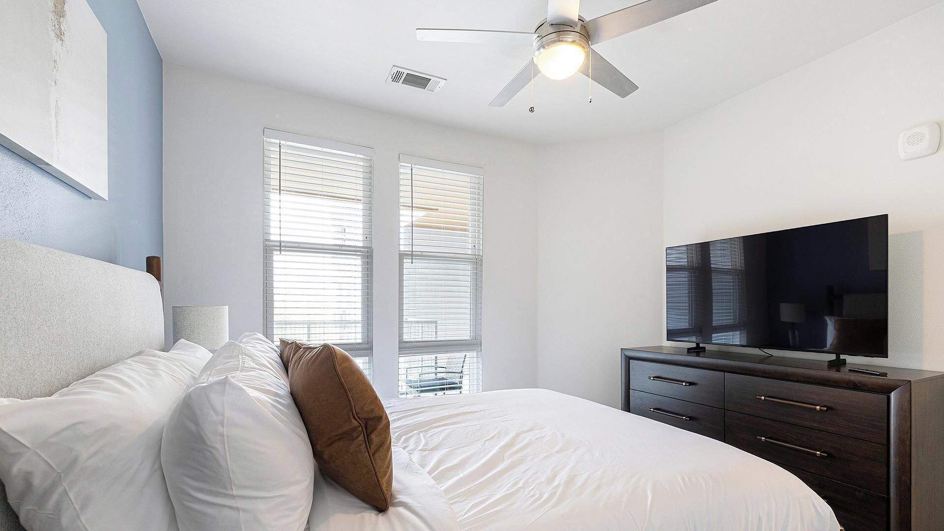 Un dormitorio con cama, ventilador de techo, televisor y dos ventanas con persianas.