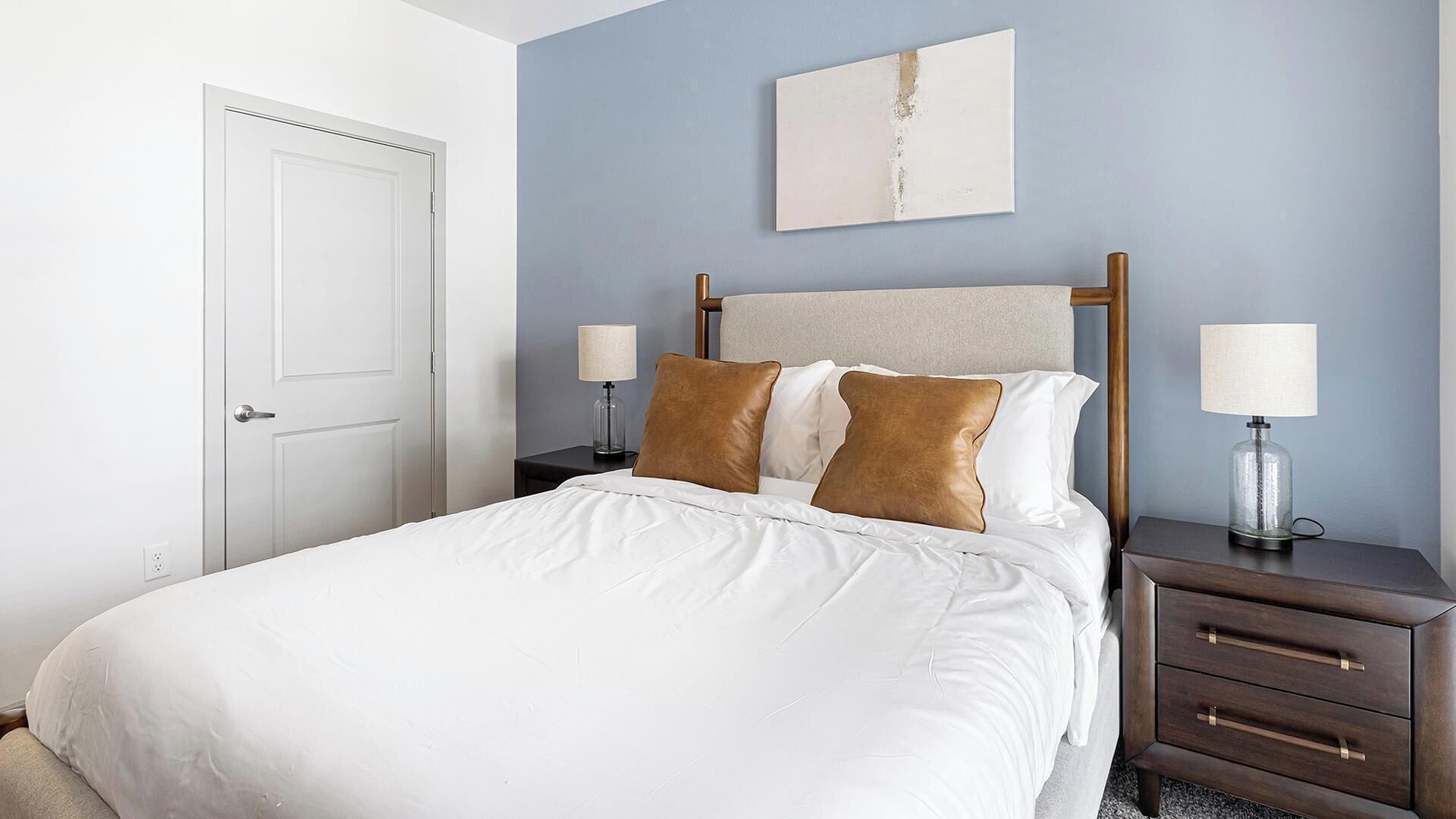 A hotel room with a bed, two bedside tables, a nightstand, a door, and a wall-mounted artwork.