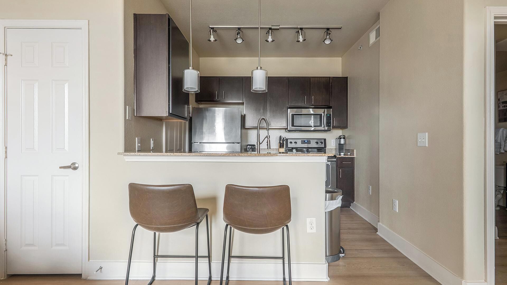 Una cocina pequeña con electrodomésticos de acero inoxidable, dos taburetes de bar e iluminación colgante.