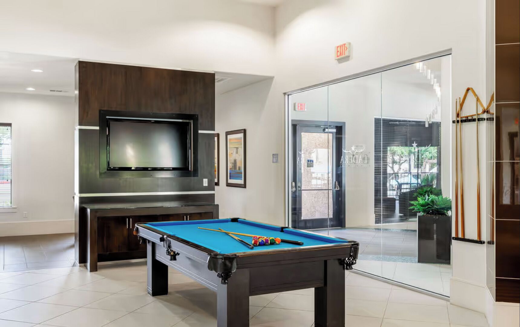 A pool table with cue sticks, a television, and a door with an exit sign.